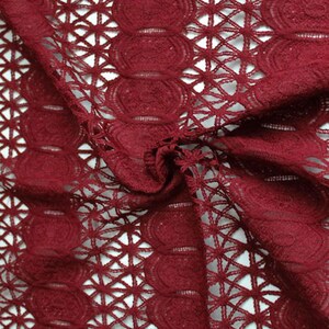 Puede incluir: Primer plano de una tela de encaje rojo oscuro con un delicado diseño calado. La tela presenta un patrón repetitivo de círculos y formas geométricas, creando una apariencia texturizada y aireada. El material está drapeado, mostrando los detalles del encaje.