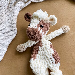 Pode incluir: Um brinquedo de pel&uacute;cia de vaca de croch&ecirc; marrom e branco com olhos pretos. O brinquedo est&aacute; deitado em uma superf&iacute;cie marrom.