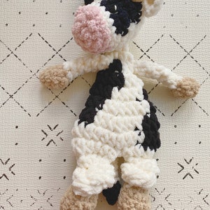 Pode incluir: Uma vaca de pel&uacute;cia de croch&ecirc; com manchas pretas e brancas, nariz rosa e orelhas marrons. A vaca est&aacute; deitada em um fundo branco com um padr&atilde;o de diamantes pretos.
