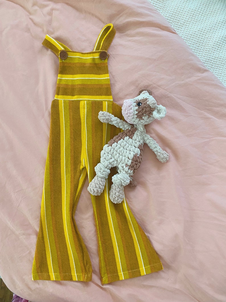 Pode incluir: Macac&atilde;o listrado amarelo e marrom com um bot&atilde;o marrom nas al&ccedil;as. Um brinquedo de pel&uacute;cia de vaca marrom e branco de croch&ecirc; est&aacute; em cima do macac&atilde;o.