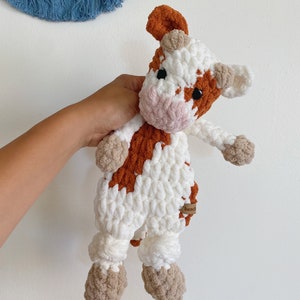Pode incluir: Um brinquedo de pel&uacute;cia de vaca crochetado com pelo marrom e branco. A vaca tem um nariz rosa e olhos pretos. O brinquedo est&aacute; sendo segurado por uma m&atilde;o. O brinquedo tem uma etiqueta marrom que diz "Beads".