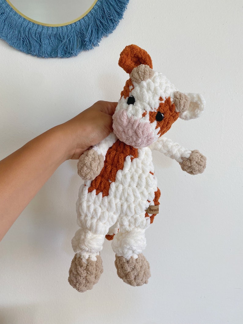 Pode incluir: Um brinquedo de pel&uacute;cia de vaca crochetado com pelo marrom e branco. A vaca tem um nariz rosa e olhos pretos. O brinquedo est&aacute; sendo segurado por uma m&atilde;o. O brinquedo tem uma etiqueta marrom que diz "Beads".