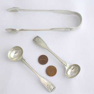 May include: A set of three silver serving pieces, including a pair of sugar tongs and a small ladle. The tongs and ladle feature a decorative shell design.