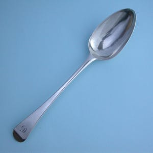 May include: A silver spoon with a long handle and an oval bowl, set against a light blue background. The spoon has a simple design with a rounded tip and a small, engraved initial on the handle.