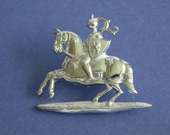 May include: A silver-toned metal pin depicting a knight on horseback. The knight is wearing armor and holding a shield with a crest. The horse is rearing up on its hind legs. The pin is mounted on a small base.