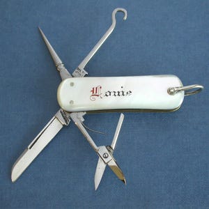 May include: A multi-tool with a mother-of-pearl handle and various implements, including a knife, scissors, and a hook. The handle is inscribed with the name "Louie" in red script. The tools are made of silver-colored metal.