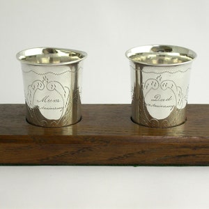 May include: Two silver cups with engraved designs and the words "Mum Anniversary" and "Dad Anniversary" on a wooden base.