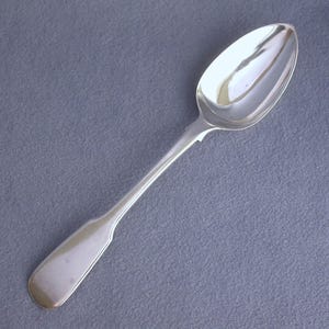 May include: A silver spoon with a rounded bowl and a long, slender handle. The spoon is highly polished, reflecting the light. The handle has a slight curve and a flat end. The spoon is set against a gray background.