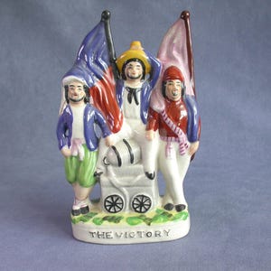 May include: A ceramic figurine depicting three figures with flags, standing near a small cart. The figures wear colorful outfits, including blue, red, and yellow. The base of the figurine reads "THE VICTORY".