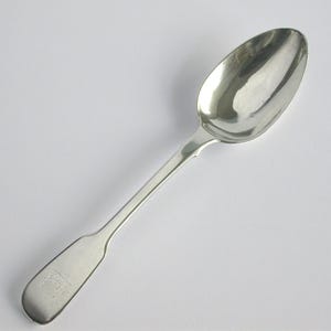 May include: A silver spoon with a rounded bowl and a long, slender handle. The handle has a rectangular end with a small, engraved design. The spoon is set against a white background.