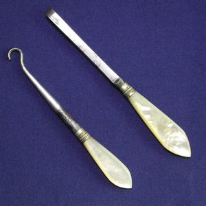 May include: Two vintage surgical tools on a blue backdrop. One is a small, curved hook, the other a flat, rectangular blade. Both have mother-of-pearl handles and metal detailing.