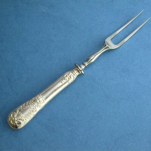 May include: Silver serving fork with two prongs and a decorative handle. The handle features ornate floral and scrollwork designs. The fork is made of a shiny, reflective metal and is set against a blue background.