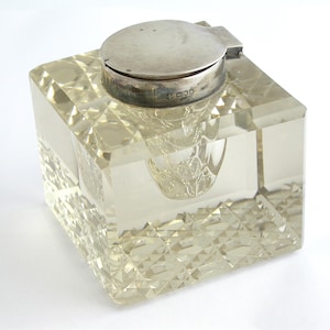 May include: A clear glass inkwell with a cut glass design and a silver lid. The lid has a small handle and a decorative inscription.