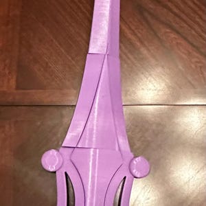 May include: A purple plastic sword with a brown wooden handle. The sword has a pointed tip and a decorative design on the hilt.