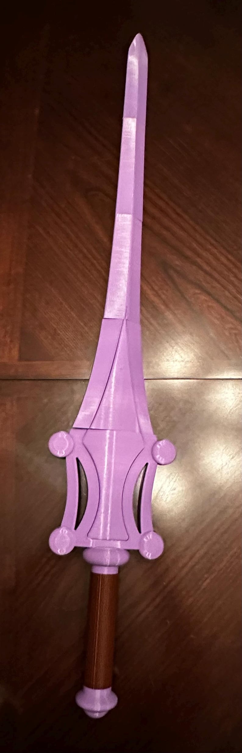 May include: A purple plastic sword with a brown wooden handle. The sword has a pointed tip and a decorative design on the hilt.
