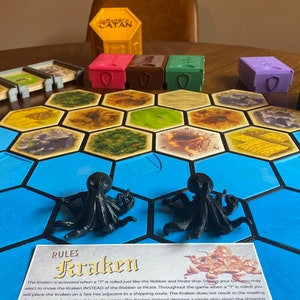 Puede incluir: Una configuración de juego de mesa con fichas hexagonales, cajas de recursos y figuras de pulpo negras. El tablero incluye fichas de agua azules y fichas de tierra con varios recursos. El juego se llama Catan e incluye una regla Kraken.