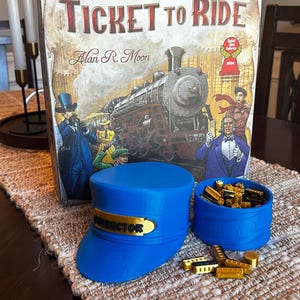 May include: A blue conductor's hat with a gold "Conductor" badge, a blue container with gold train car pieces, and a board game box with the title "Ticket to Ride" by Alan R. Moon.