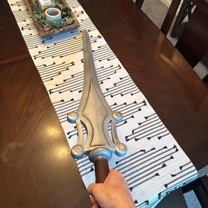 May include: A silver-colored sword with a brown handle. The sword has a decorative design with multiple circular accents. The sword is being held by a hand.
