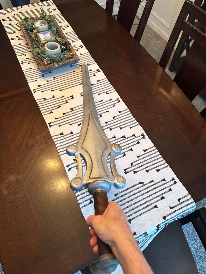 May include: A silver-colored sword with a brown handle. The sword has a decorative design with multiple circular accents. The sword is being held by a hand.
