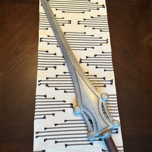May include: A silver-colored sword with a decorative handle and a pointed blade. The sword is lying on a white and black striped fabric.
