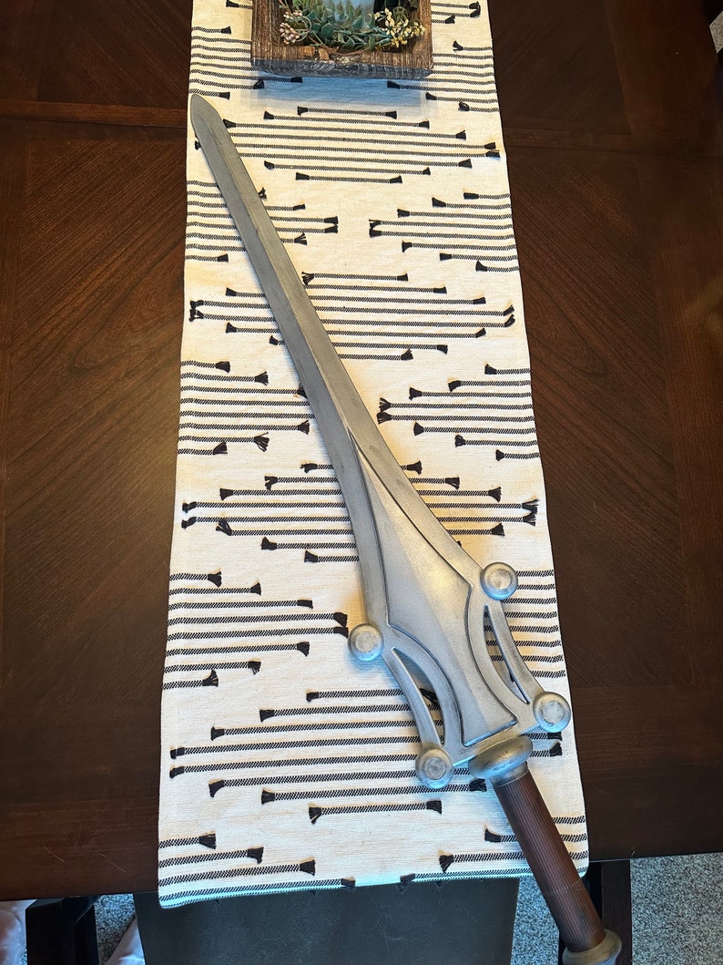 May include: A silver-colored sword with a decorative handle and a pointed blade. The sword is lying on a white and black striped fabric.