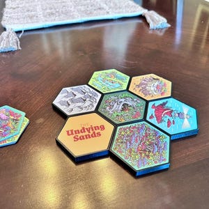 May include: A collection of colorful, hexagonal game tiles with various illustrations and the text "The Undying Sands." The tiles are arranged in a flower-like pattern on a dark wooden surface. One set of tiles is separate from the main group.