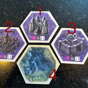 Puede incluir: Cuatro fichas hexagonales de juego con ilustraciones de castillos y un punto rosa y dados en cada ficha. Las fichas están numeradas del 1 al 4.