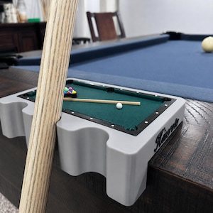 May include: A miniature pool table accessory in light gray plastic with a green felt surface, featuring a small cue stick, a rack of tiny billiard balls, and a white cue ball. The accessory includes a holder for a full-sized pool cue stick.