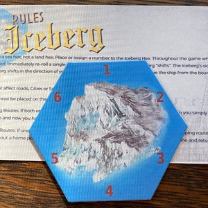 Puede incluir: Una pieza de juego hexagonal azul con una imagen blanca y gris de un iceberg. Los números del 1 al 6 están impresos en los lados de la pieza en rojo.