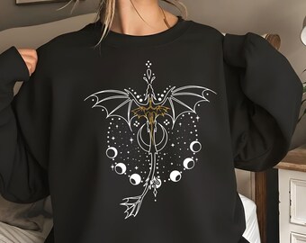 May include: A black crewneck sweatshirt featuring a central graphic of a stylized dragon with wings spread wide. The design includes white celestial elements, including a crescent moon and a series of moon phases, surrounded by small white dots.