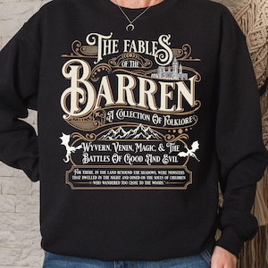 May include: Black sweatshirt with a cream-colored graphic. The graphic reads "The Fables of the Barren: A Collection of Folklore" with additional text about wyverns, magic, and battles. A castle and mountains are also depicted.