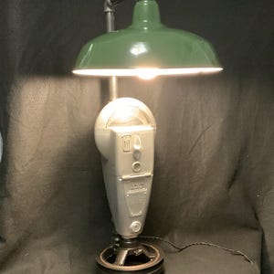 Puede incluir: Una lámpara de escritorio única hecha de materiales reciclados. Cuenta con una pantalla de metal verde, un cuerpo de parquímetro plateado y una base de polea. La lámpara tiene un cable negro y una estética industrial vintage.