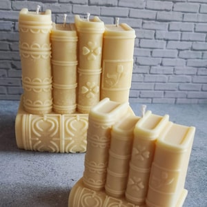 May include: Two ivory-colored candles shaped like stacks of books. Each candle features detailed embossed designs, including floral and geometric patterns. The wicks are visible at the top of each candle, suggesting they are ready to be lit.