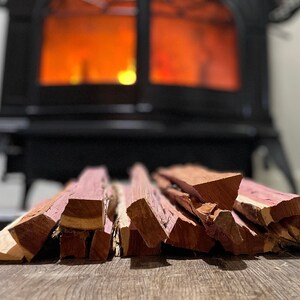 May include: A close-up of a pile of reddish-brown wood chips, likely for smoking meat, with a wood-burning stove in the background.
