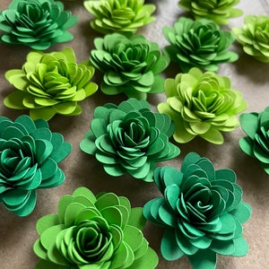 May include: A collection of 14 green paper flowers, each with multiple layers of petals, arranged in a random pattern on a brown surface.