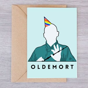 Puede incluir: Una tarjeta de felicitación azul claro con una ilustración de dibujos animados de Lord Voldemort con un gorro de fiesta arcoíris y sosteniendo una varita mágica. El texto "OLDEMORT" está impreso en negro en la parte inferior de la tarjeta.