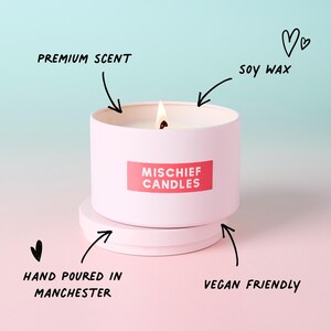May include: A light pink metal candle tin with a lit wick and a matching lid underneath. A red rectangular label features the white text MISCHIEF CANDLES. Hand-drawn arrows point to the text labels: PREMIUM SCENT, SOY WAX, HAND POURED IN MANCHESTER, and VEGAN FRIENDLY.