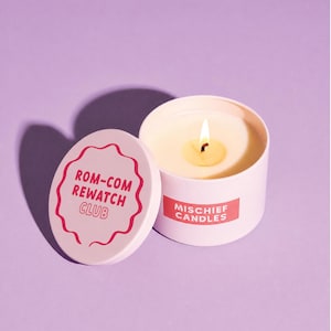 May include: A light pink metal candle tin with a lit white wax candle inside. The lid is placed beside the tin, featuring a wavy red border and the text ROM-COM REWATCH CLUB. A red rectangular label on the tin reads MISCHIEF CANDLES in white.