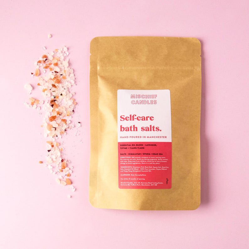 May include: A brown paper pouch of bath salts with a white and red label reading Mischief Candles Self-care bath salts. Hand-poured in Manchester. A pile of pink and white salt crystals sits to the left of the pouch on a light pink surface.
