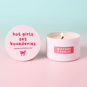 May include: A light pink candle in a round metal tin with a lit wick. The lid is placed to the left, featuring the dark pink text "hot girls set boundaries" and a small bow graphic. The side of the tin displays a red label with "MISCHIEF CANDLES".