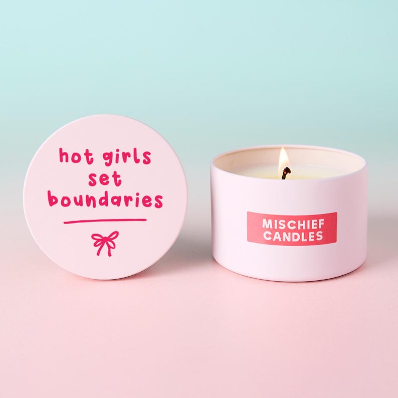 May include: A light pink candle in a round metal tin with a lit wick. The lid is placed to the left, featuring the dark pink text "hot girls set boundaries" and a small bow graphic. The side of the tin displays a red label with "MISCHIEF CANDLES".