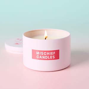May include: A light pink cylindrical candle tin with a lit wick and a white wax interior. A rectangular red label on the front features the white text "MISCHIEF CANDLES" in a bold, sans-serif font. A matching pink lid rests to the left of the candle.