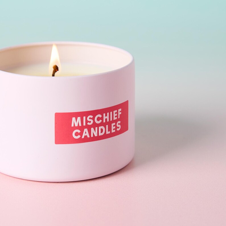 May include: A light pink cylindrical candle tin with a lit wick and a red rectangular label featuring the white text MISCHIEF CANDLES. The candle is filled with white wax and the container has a smooth, matte finish on the exterior surface.