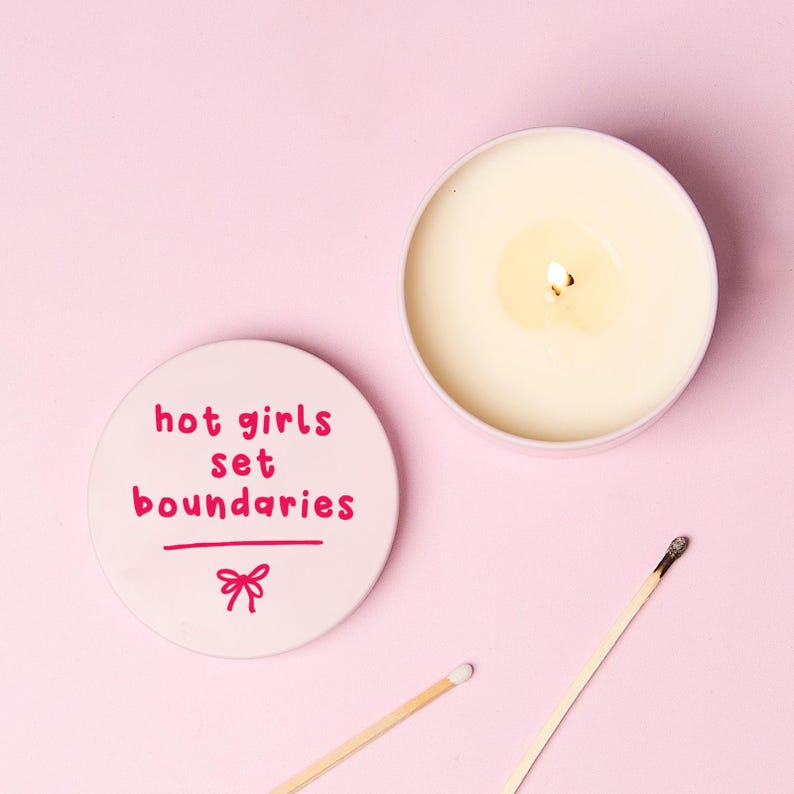 May include: A round pink candle tin with a lid featuring the text "hot girls set boundaries" in dark pink lettering above a small bow icon. The candle is lit, showing white wax, and is accompanied by two wooden matches on a pink surface.