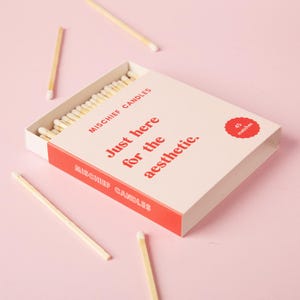 May include: A rectangular matchbox with a light pink cover and a red side panel. The text reads MISCHIEF CANDLES and Just here for the aesthetic. A red circular badge indicates 45 matches. Several wooden matches with white tips are scattered around.