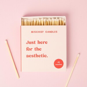 May include: A rectangular matchbox with a light pink cover featuring the text MISCHIEF CANDLES and Just here for the aesthetic. in red font. A red scalloped circle indicates 45 matches. The box is open, revealing wooden matches with white tips.