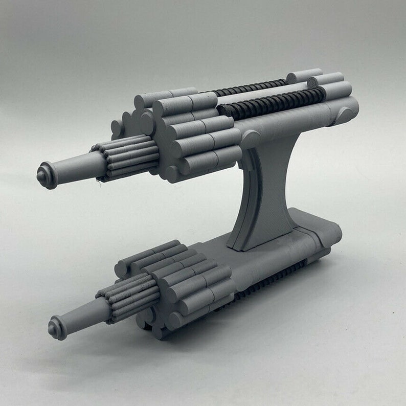 May include: A gray 3D printed model of a double-barreled weapon with a futuristic design. The weapon has a cylindrical body with a series of small, cylindrical protrusions along the sides. The weapon is mounted on a base with a U-shaped design.
