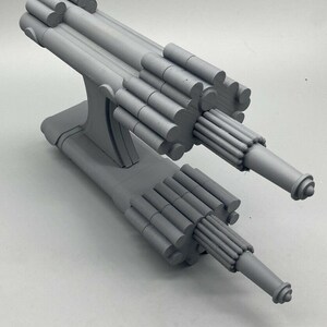May include: A gray 3D printed model of a futuristic space ship with a long, cylindrical body and two cannons on the front.