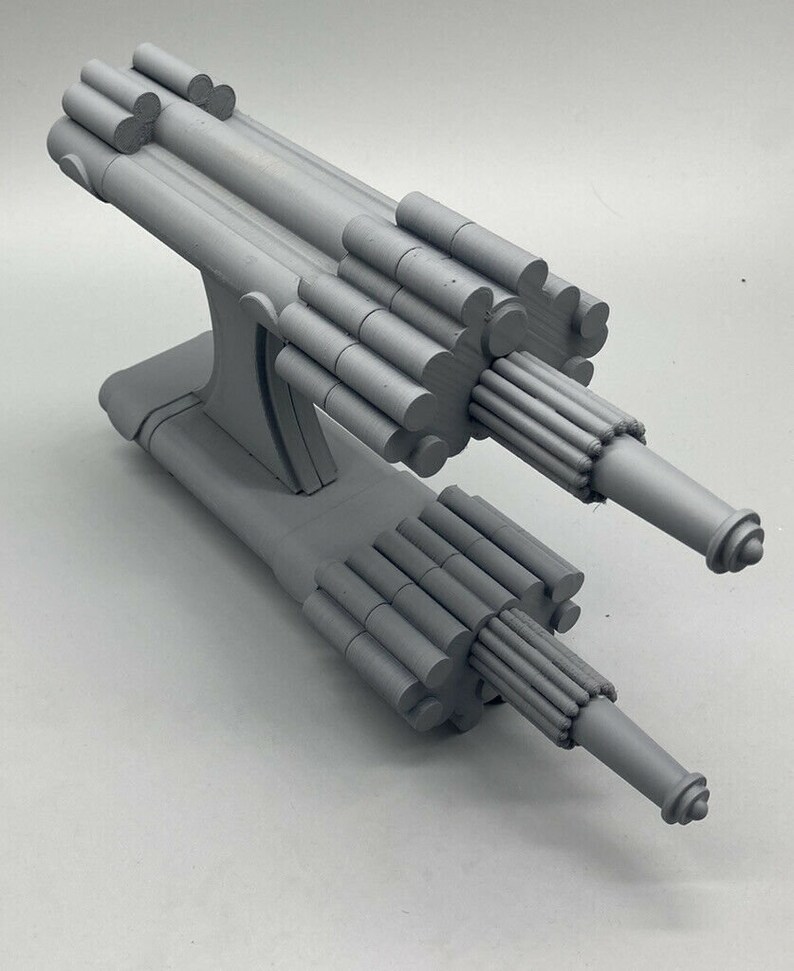 May include: A gray 3D printed model of a futuristic space ship with a long, cylindrical body and two cannons on the front.