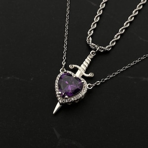 May include: A silver necklace featuring a heart-shaped pendant with a purple gemstone center and a small dagger. The pendant is surrounded by small, clear stones. The necklace is on a black surface.